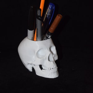 May include: White skull-shaped desk organizer with a pencil, pen, and two markers sticking out of the top.