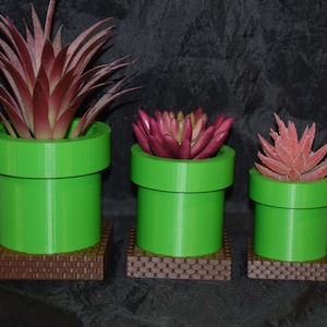 May include: Three green and brown ceramic planters shaped like a Super Mario Bros. pipe, each with a different type of artificial succulent plant.