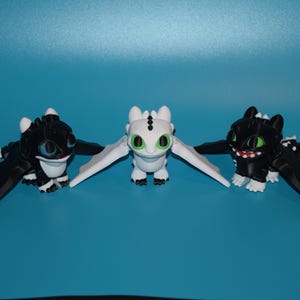 May include: Three dragon figurines in a row, each with outstretched wings. Two are black with white accents, and one is white. The dragons have detailed features, including eyes, teeth, and claws. The background is a solid blue.
