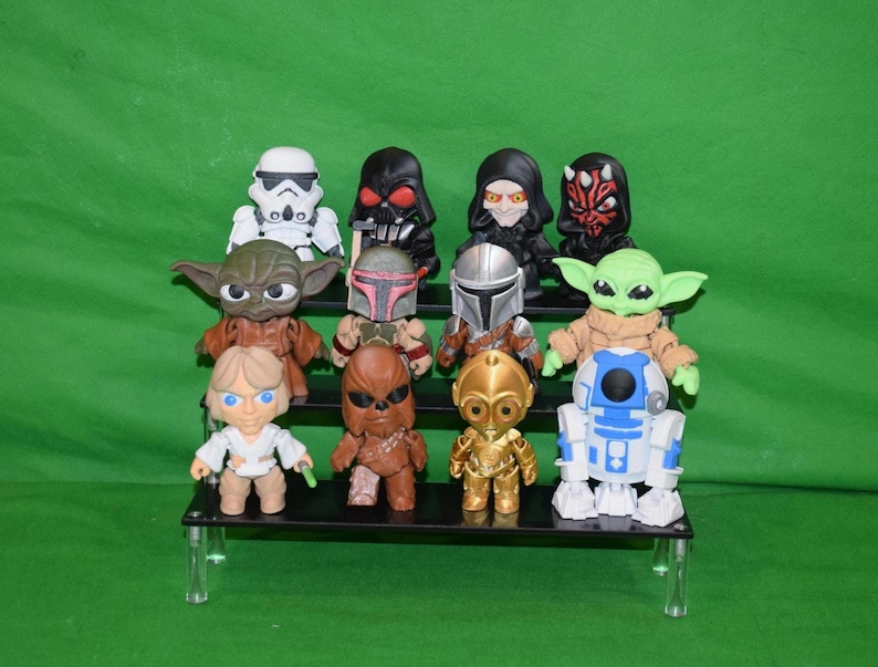 Op de afbeelding: Een verzameling Star Wars-personagefiguren, waaronder Darth Vader, Yoda en R2-D2, tentoongesteld op een zwarte trapvormige plank. De beeldjes zijn felgekleurd en gedetailleerd, met een groene achtergrond.