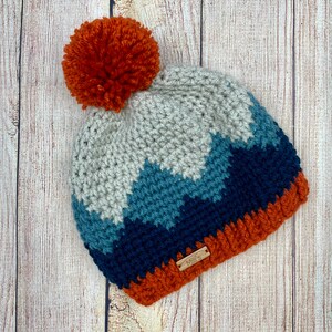 K&ouml;nnte beinhalten: Eine geh&auml;kelte M&uuml;tze mit einem Bommel oben. Die M&uuml;tze ist in einem Chevron-Muster mit Blaut&ouml;nen, Grau und Orange gearbeitet. Die M&uuml;tze hat ein kleines braunes Etikett mit der Aufschrift "Mimi's".