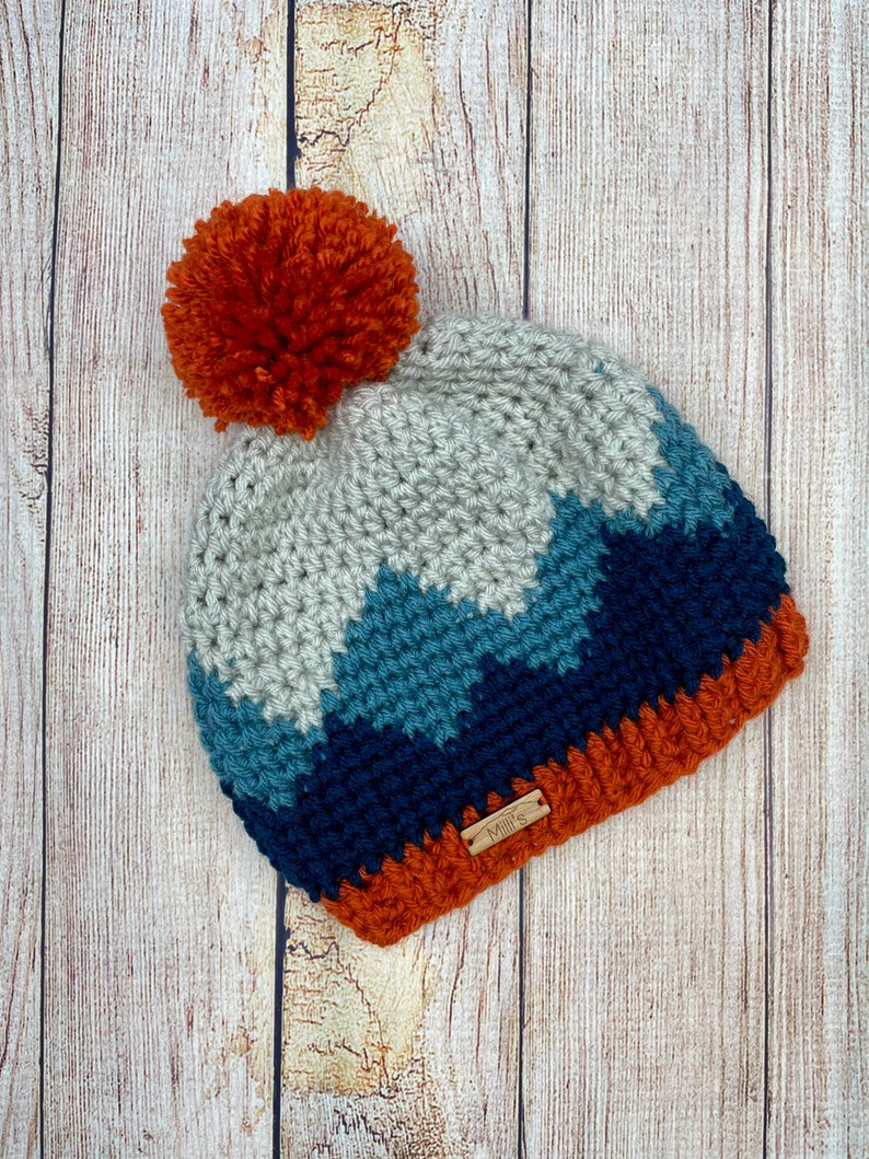 K&ouml;nnte beinhalten: Eine geh&auml;kelte M&uuml;tze mit einem Bommel oben. Die M&uuml;tze ist in einem Chevron-Muster mit Blaut&ouml;nen, Grau und Orange gearbeitet. Die M&uuml;tze hat ein kleines braunes Etikett mit der Aufschrift "Mimi's".