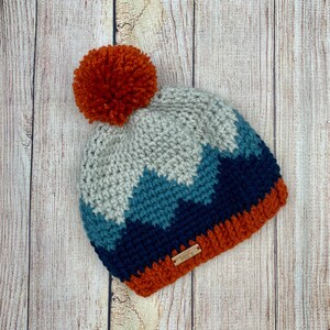 K&ouml;nnte beinhalten: Eine geh&auml;kelte M&uuml;tze mit einem Bommel oben. Die M&uuml;tze ist in einem Chevron-Muster mit Blaut&ouml;nen, Grau und Orange gearbeitet. Die M&uuml;tze hat ein kleines braunes Etikett mit der Aufschrift "Annie's".