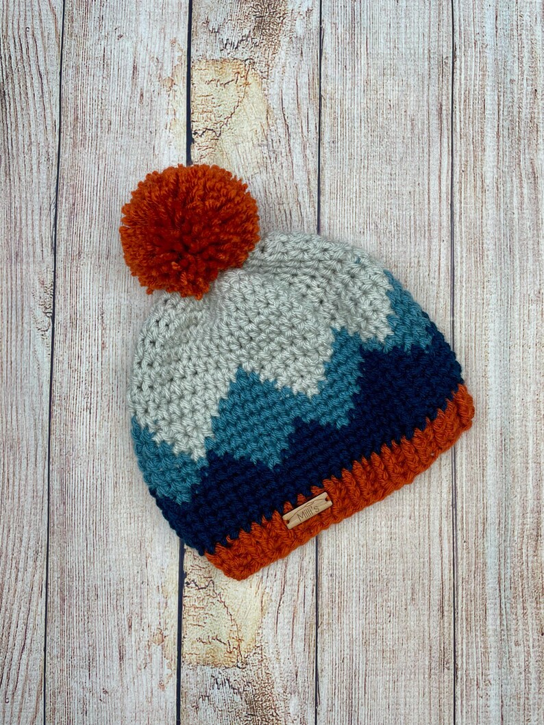 K&ouml;nnte beinhalten: Eine geh&auml;kelte M&uuml;tze mit einem Bommel oben. Die M&uuml;tze ist in einem Chevron-Muster mit Blaut&ouml;nen, Grau und Orange gearbeitet. Die M&uuml;tze hat ein kleines braunes Etikett mit der Aufschrift "Annie's".