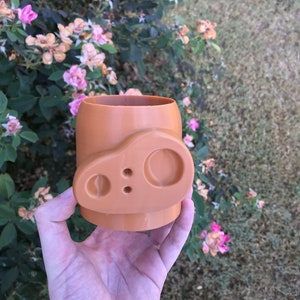 May include: A terracotta-colored ceramic planter with a unique design featuring two rounded holes and a smaller hole in the center. The planter is held in a hand.