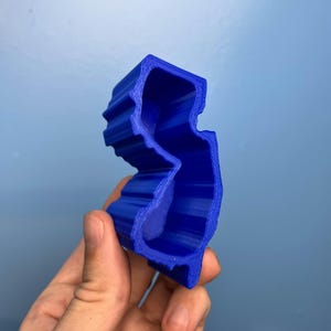 May include: A 3D-printed blue cookie cutter in the shape of the state of New Jersey. The cutter is held in a hand against a light blue background. The cutter has a ridged exterior.