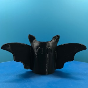 Peut inclure: Un porte-crayons en forme de chauve-souris imprim&eacute; en 3D noir avec deux trous pour les crayons. Le porte-crayons est pos&eacute; sur une surface bleue.