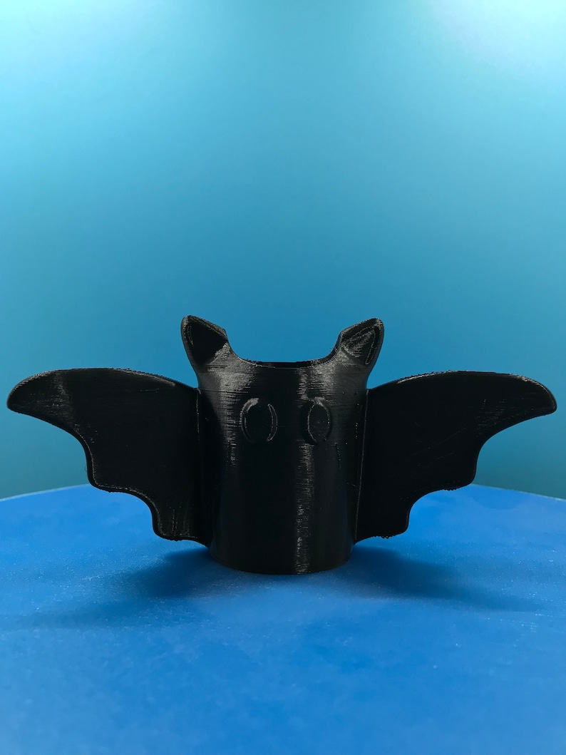 Peut inclure: Un porte-crayons en forme de chauve-souris imprim&eacute; en 3D noir avec deux trous pour les crayons. Le porte-crayons est pos&eacute; sur une surface bleue.