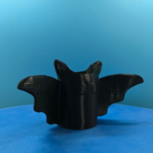 Peut inclure: Un porte-crayons en forme de chauve-souris imprim&eacute; en 3D de couleur noire. Le porte-crayons a deux petits cercles pour les yeux et une large envergure.