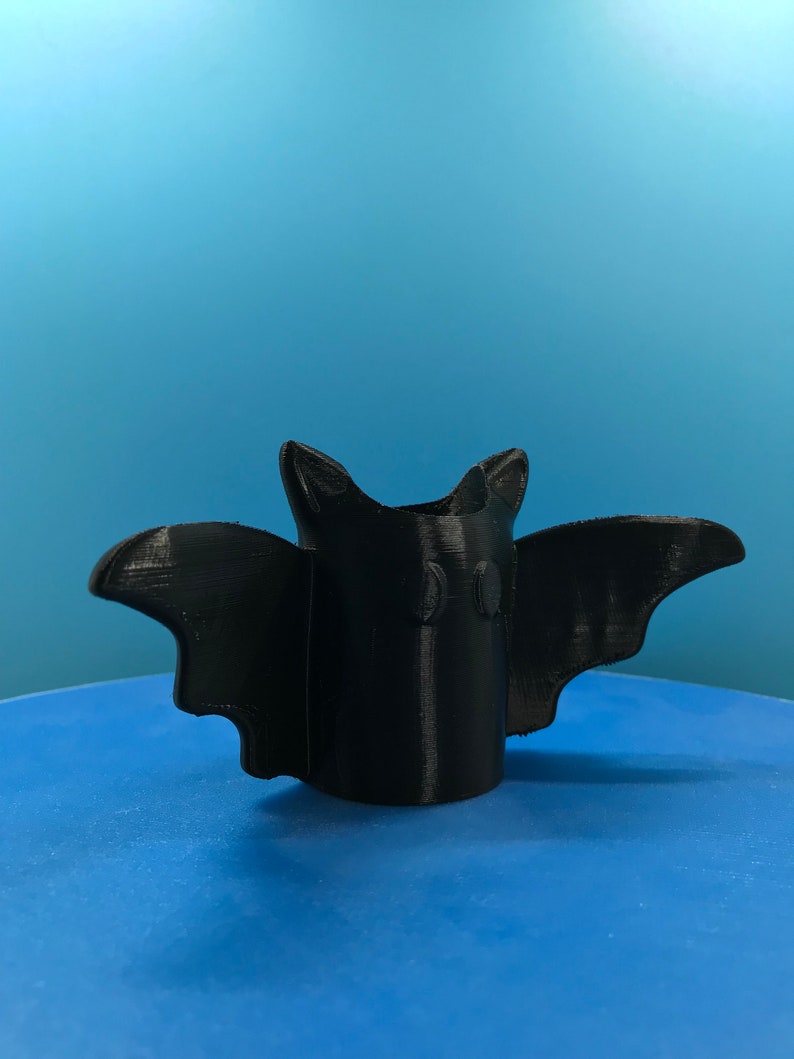 Peut inclure: Un porte-crayons en forme de chauve-souris imprim&eacute; en 3D de couleur noire. Le porte-crayons a deux petits cercles pour les yeux et une large envergure.