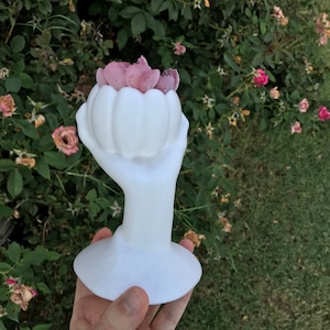 May include: A white ceramic sculpture of a hand holding a flower-shaped vase filled with pink petals. The sculpture has a smooth, glossy finish and sits on a round base. The background features green foliage and a few pink flowers.