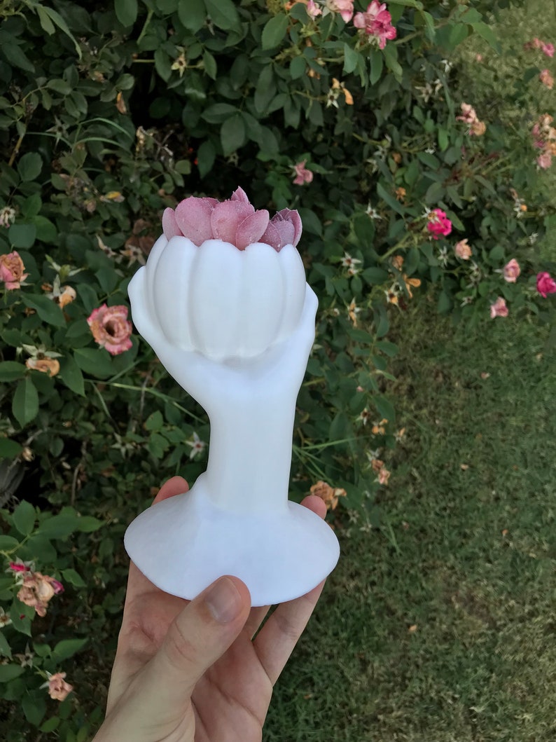 May include: A white ceramic sculpture of a hand holding a flower-shaped vase filled with pink petals. The sculpture has a smooth, glossy finish and sits on a round base. The background features green foliage and a few pink flowers.