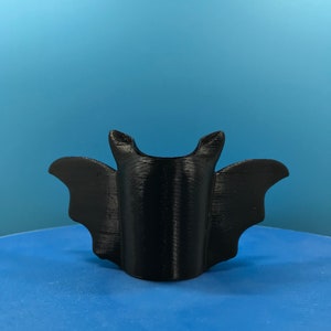 Peut inclure: Un support de t&eacute;l&eacute;phone en forme de chauve-souris imprim&eacute; en 3D noir. Le support est con&ccedil;u pour maintenir un t&eacute;l&eacute;phone en position verticale.