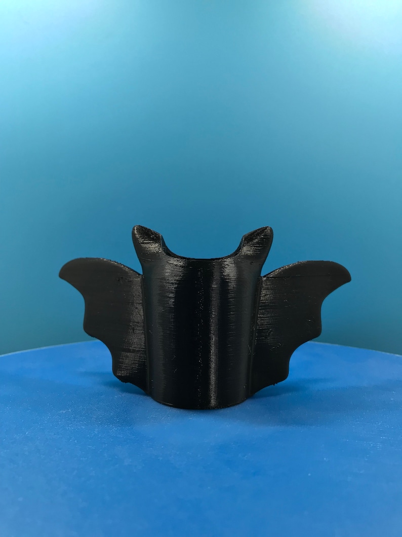 Peut inclure: Un support de t&eacute;l&eacute;phone en forme de chauve-souris imprim&eacute; en 3D noir. Le support est con&ccedil;u pour maintenir un t&eacute;l&eacute;phone en position verticale.