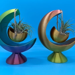May include: Two crescent-shaped air plant holders in a gradient of colors, one with green, blue, and purple, the other with gold, orange, and green. Each holds an air plant in a gold-colored bowl suspended by twine. The bases are round.