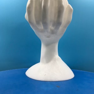 May include: A white sculpture of a hand with fingers curled inwards, creating a cup-like form. It stands on a thin neck and a flared base. The background is a solid blue.