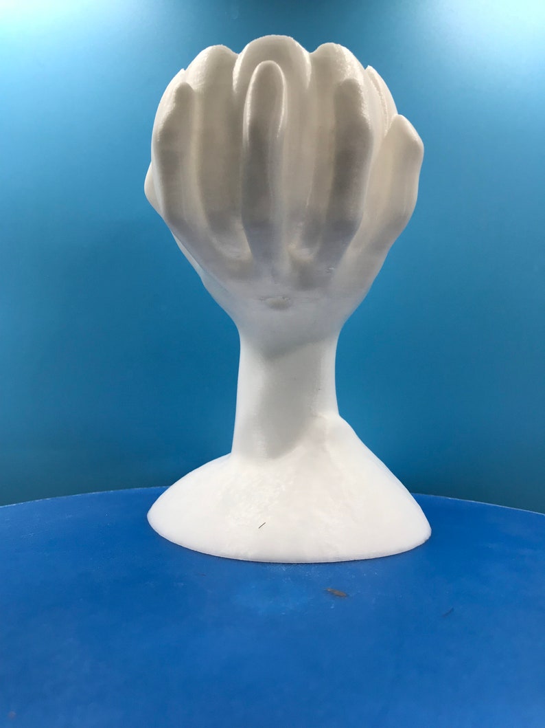 May include: A white sculpture of a hand with fingers curled inwards, creating a cup-like form. It stands on a thin neck and a flared base. The background is a solid blue.