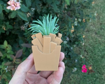 May include: A tan-colored planter shaped like a french fries container, filled with a small, green succulent. The planter is held in a hand against a backdrop of green foliage and pink flowers. The planter is a unique decorative item.
