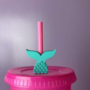 May include: A pink plastic tumbler with a matching lid. A teal mermaid tail straw topper is inserted into a pink straw. The background is a solid light purple.