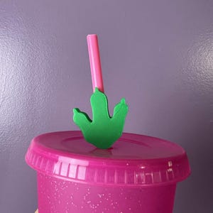 May include: A bright pink glittery tumbler with a matching lid and a pink straw. Attached to the straw is a green, three-toed dinosaur footprint topper. The cup has a textured, sparkling finish and a ridged lid design, creating a vibrant look.