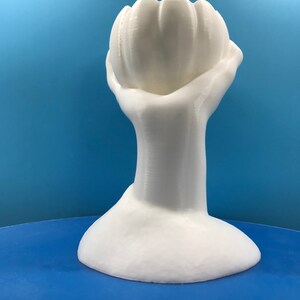 May include: A white, 3D-printed sculpture of a hand holding a flower-like shape. The sculpture has a smooth, matte finish and sits on a round base. The background is a solid blue.