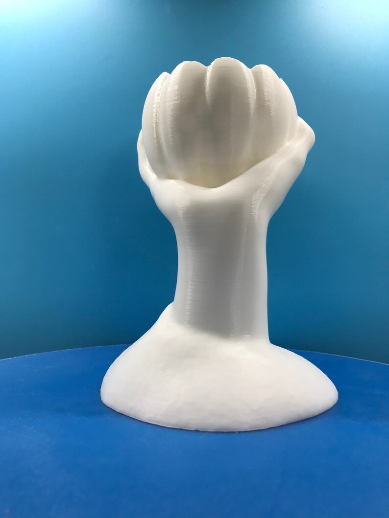 May include: A white, 3D-printed sculpture of a hand holding a flower-like shape. The sculpture has a smooth, matte finish and sits on a round base. The background is a solid blue.