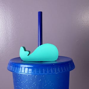 May include: A bright turquoise whale-shaped straw topper attached to a dark blue straw. The straw is inserted into a dark blue, glitter-flecked tumbler lid. The whale topper is a flat, solid-colored cutout that sits horizontally on the lid.
