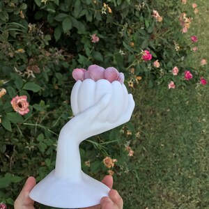 May include: A white ceramic sculpture of a hand holding a flower-shaped vase filled with pink, textured spheres. The sculpture has a wide, flat base and is set against a backdrop of green foliage and pink roses.