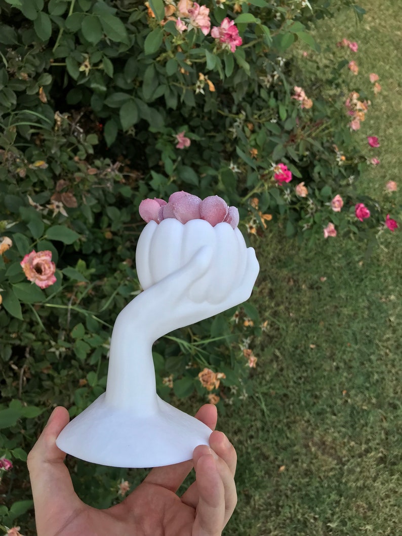 May include: A white ceramic sculpture of a hand holding a flower-shaped vase filled with pink, textured spheres. The sculpture has a wide, flat base and is set against a backdrop of green foliage and pink roses.