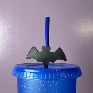 May include: A blue plastic tumbler with a matching lid and a blue straw. A black, flat, bat-shaped topper is attached to the straw, resting just above the lid. The tumbler features a translucent finish with small, embedded white glitter particles.