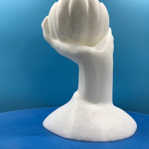 May include: A white sculpture of a hand holding a stylized flower. The hand and flower are smooth and rounded, with subtle texture. The base is a rounded shape. The background is a gradient of blue.