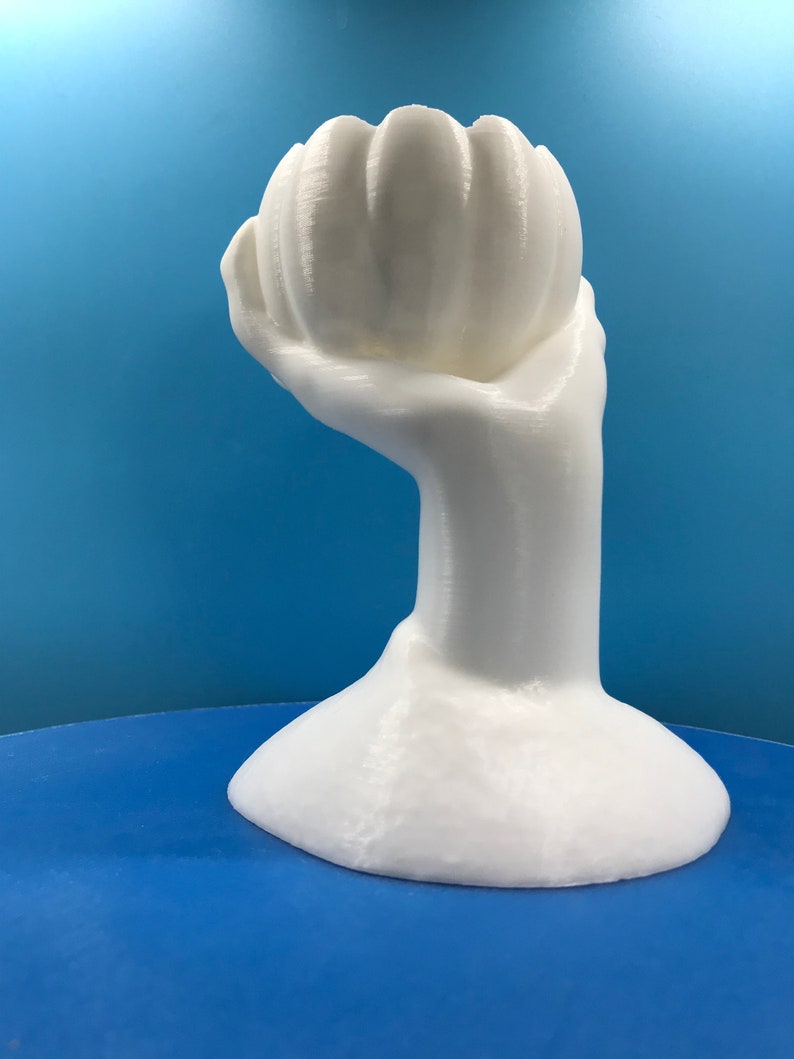 May include: A white sculpture of a hand holding a stylized flower. The hand and flower are smooth and rounded, with subtle texture. The base is a rounded shape. The background is a gradient of blue.