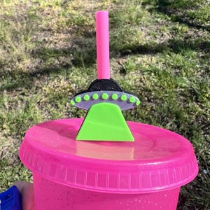 May include: A pink reusable cup with a black and green UFO straw topper. The UFO has a green top and a black base with silver accents.