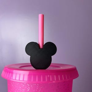 May include: A pink plastic tumbler with a matching lid and a black Mickey Mouse-shaped straw topper. A pink straw is inserted into the topper. The tumbler has a glittery appearance and sits against a purple background.