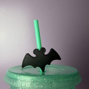 May include: A black 3D-printed bat-shaped straw topper attached to a light green plastic straw. The straw is inserted into the lid of a translucent light green tumbler that features embedded silver glitter throughout the plastic material.