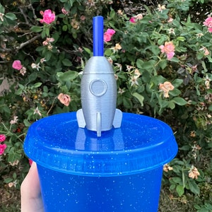May include: A blue glitter tumbler with a silver rocket ship straw topper. The rocket ship has a blue straw and a silver body with a round window.