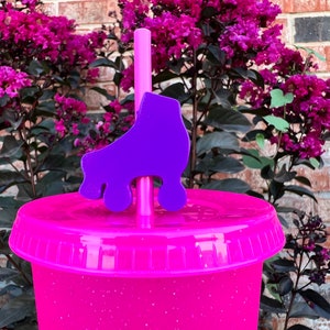 May include: A pink glitter tumbler with a purple roller skate straw topper. The tumbler is sitting on a table with a purple flower bush in the background.