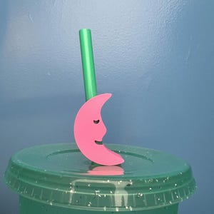 May include: A pink crescent moon straw topper with a smiling face, resting on a green glittery cup lid. A green straw is inserted into the lid. The background is a solid blue.