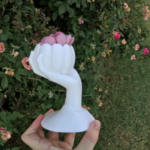 May include: A white, hand-shaped decorative vase holding a cluster of pink flowers. The vase has a rounded base and a slender, curved stem that forms the hand. The flowers are a soft pink colour, contrasting with the white vase. The background is a green bush.