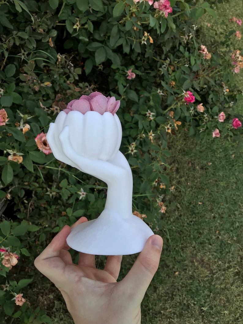 May include: A white, hand-shaped decorative vase holding a cluster of pink flowers. The vase has a rounded base and a slender, curved stem that forms the hand. The flowers are a soft pink colour, contrasting with the white vase. The background is a green bush.