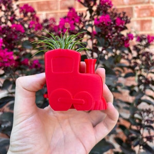 May include: A red plastic train planter with a small air plant growing inside. The planter is being held in a hand.