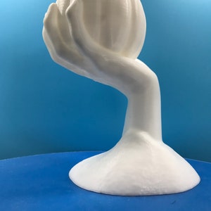 May include: A white sculpture depicting a hand holding a flower-like shape. The artwork has a smooth finish and a rounded base. The background is a gradient of blue.
