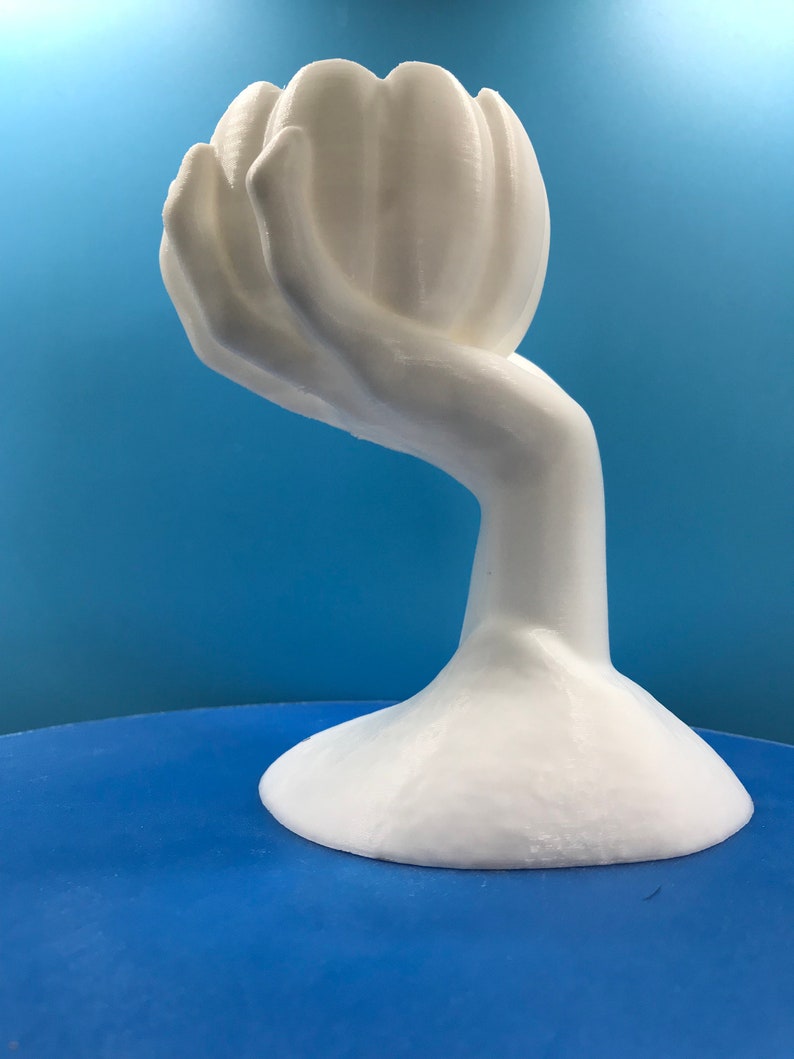 May include: A white sculpture depicting a hand holding a flower-like shape. The artwork has a smooth finish and a rounded base. The background is a gradient of blue.