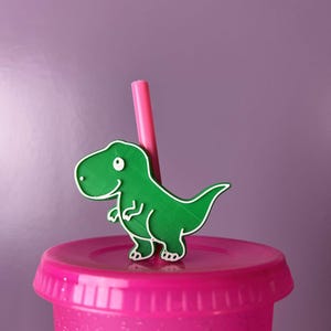 May include: A bright green T-Rex dinosaur straw topper attached to a pink straw, which is inserted into a matching pink plastic tumbler lid. The dinosaur figure features a white outline and a single white eye, providing a clear contrast.