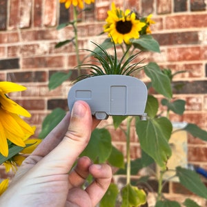 May include: A gray ceramic planter shaped like a vintage camper with a small green plant growing inside. The planter is being held in a hand against a brick wall background with sunflowers growing in front of the wall.