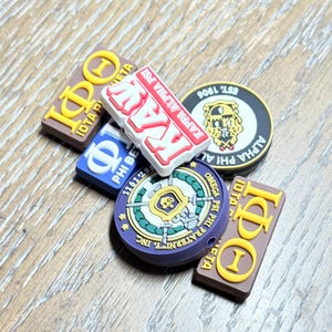 May include: A collection of colorful shoe charms featuring Greek letters and fraternity/sorority insignia. The charms are in various shapes and colors, including brown, white, blue, and purple, with gold and yellow accents.
