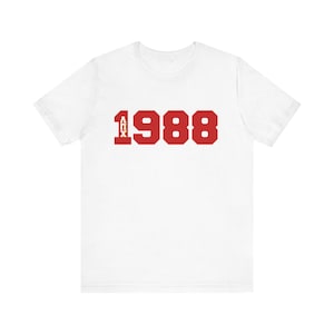 May include: White t-shirt featuring the red text "1988" in a bold, blocky font. Above the "1988" is the text "AIX" in a smaller, vertical font. The shirt is made of a soft material and has short sleeves.