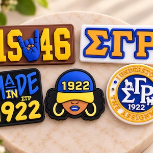 May include: Five decorative PVC shoe charms featuring blue and yellow designs. One rectangular charm reads 1946 with a hand sign, one reads ΣΓΡ, one reads MADE IN 1922, one shows a person wearing a 1922 cap, and one is a circular ΣΓΡ 1922 emblem.