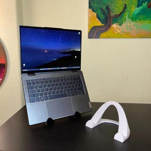 Könnte beinhalten: Ein silberner Laptop mit beleuchtetem Bildschirm, der eine Nachthimmel-Szene zeigt, wird von einem schwarzen Ständer gehalten. Ein weißer Laptop-Ständer ist ebenfalls vorhanden. Der Laptop steht auf einem dunkelbraunen Schreibtisch.