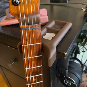 Könnte beinhalten: Nahaufnahme eines Gitarrenhalses aus Holz mit silbernen Bünden und Saiten. Ein brauner Plektrumhalter mit Plektren ist an einer dunklen Oberfläche befestigt. Ein schwarzer Kopfhörer ist zu sehen.